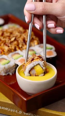 Sushi à emporter plongé dans une sauce jaune avec des baguettes, plateau de plusieurs makis en arrière-plan.