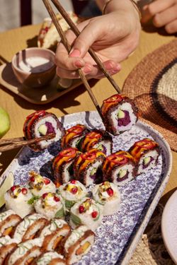 Sushis variés servis sur un plateau, avec une main utilisant des baguettes pour les prendre.