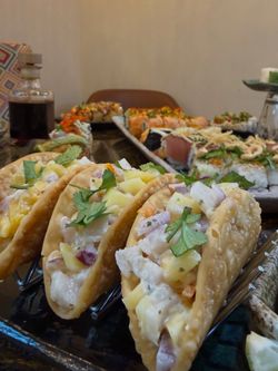 Sushi à emporter avec des tacos farcis de poisson, garnis de coriandre, accompagnés de plats de sushi variés en arrière-plan.