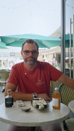 Homme portant un t-shirt Coté Sushi Tours, assis à une table avec un plateau de sushi à emporter et boissons.