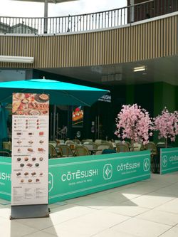 Terrasse extérieure d'un restaurant de sushis avec parasol turquoise, menu affiché et décorations florales roses. Tables et c