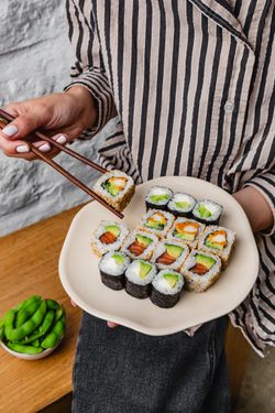 Sushi à emporter variés présentés sur une assiette tenue par une personne avec des baguettes. Edamame en accompagnement.