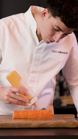 Chef en tenue blanche préparant un sushi avec un rouleau de saumon sur une planche en bois.