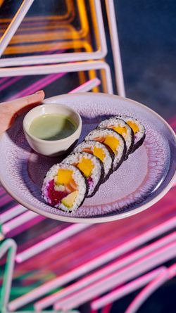Rouleau de sushi coloré avec une petite coupelle de sauce, présenté sur une assiette ronde texturée.