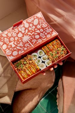 Boîte de sushi à emporter avec rouleaux et garnitures variées tenue en main, prête à la livraison.
