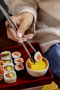 Sushi à emporter avec rouleaux variés et sauce jaune, tenu avec des baguettes dans un plateau rouge