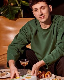 Homme en sweat vert préparant des plateaux de sushi à emporter sur une table avec un verre à pied rempli de boisson.