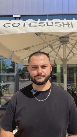 Homme avec barbe debout devant un parasol marqué COTE SUSHI à l'entrée du restaurant japonais Côté Sushi Plan de Campagne.
