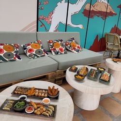 Sushi variés et plats japonais sur tables basses devant un canapé avec coussins colorés et mur décoré d'une peinture murale.