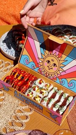 Boîte colorée ouverte contenant des sushis variés, avec des rouleaux sur une assiette en arrière-plan.