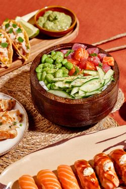Bol en bois avec sushi à emporter, edamame, concombre, saumon et thon, avec accompagnements variés en arrière-plan.