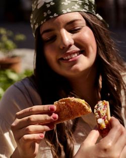 Une femme souriante tenant deux tacos garnis de légumes et fromage, dégustant un moment de repas en extérieur.