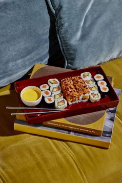 Plateau de sushi variés avec sauce jaune, présenté sur une tablette rouge, posé sur des livres sur un canapé jaune.