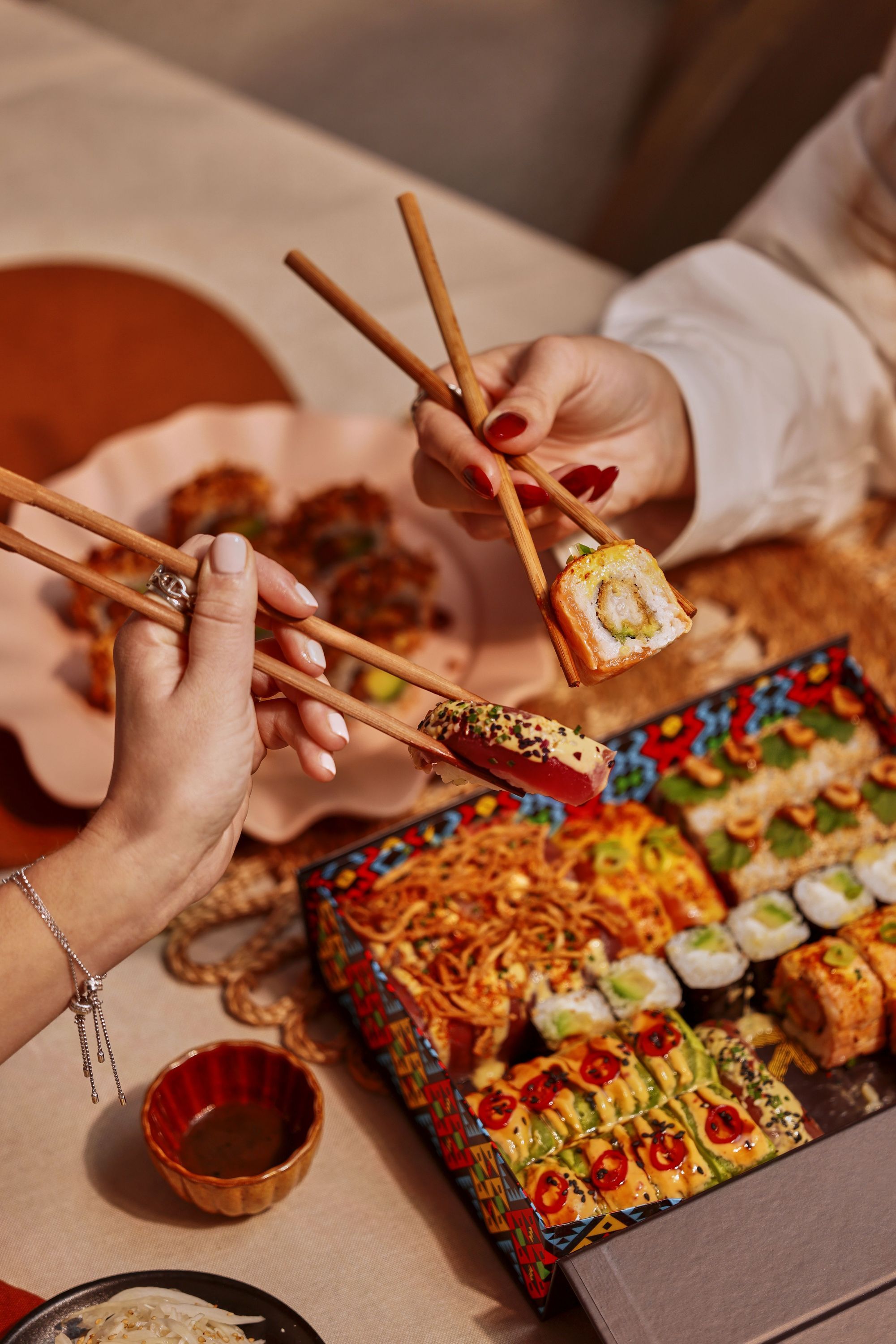 Assortiment coloré de sushis variés servis dans une boîte, avec deux personnes utilisant des baguettes pour les prendre.