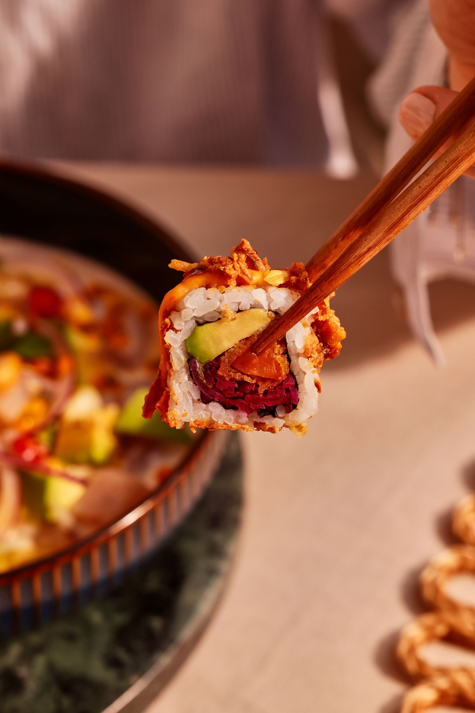 Rouleau de sushi tenu par des baguettes avec avocat, riz et garniture croustillante dans un restaurant japonais.