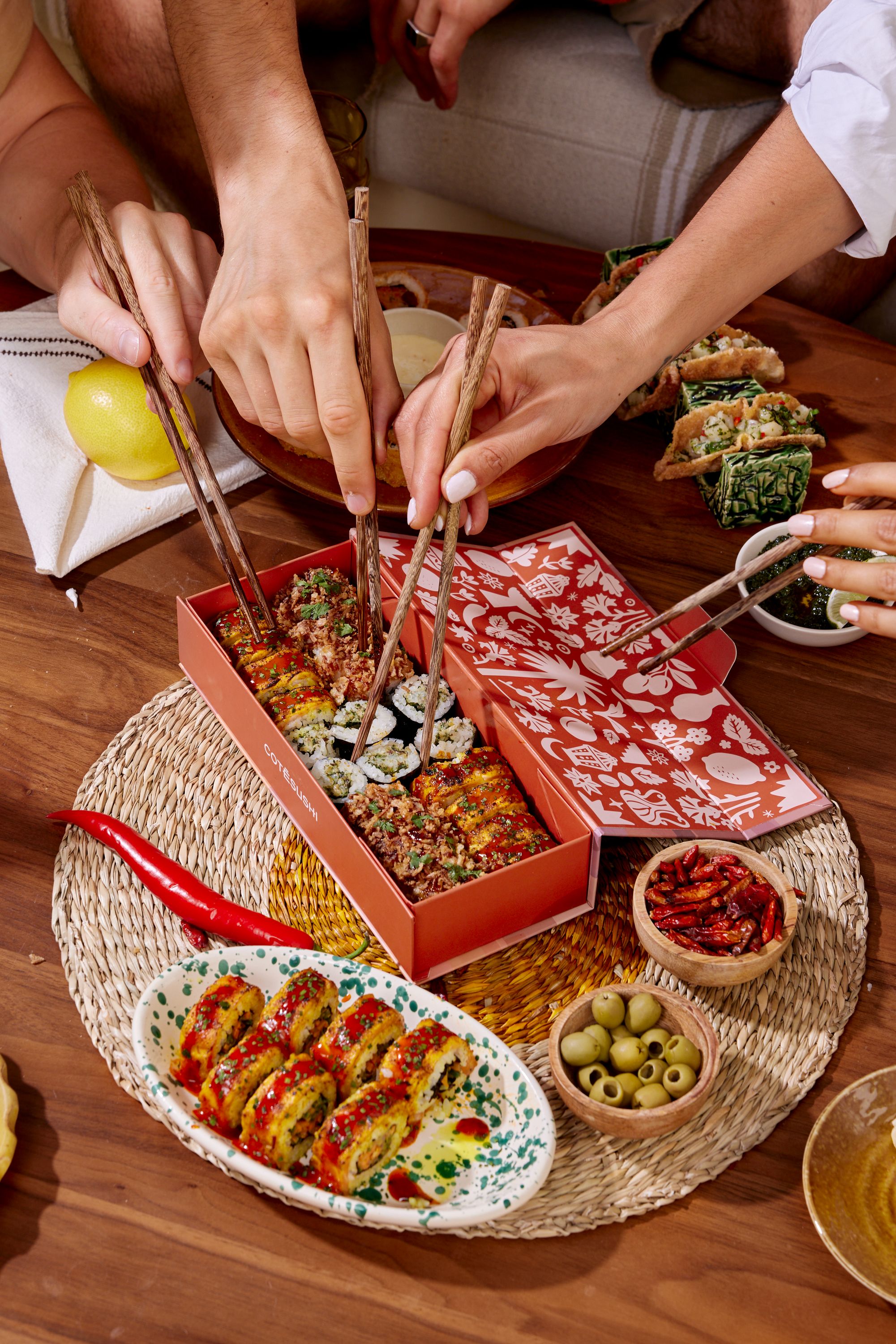 Sushi à emporter présenté dans une boîte décorée, avec plusieurs mains utilisant des baguettes pour manger ensemble.