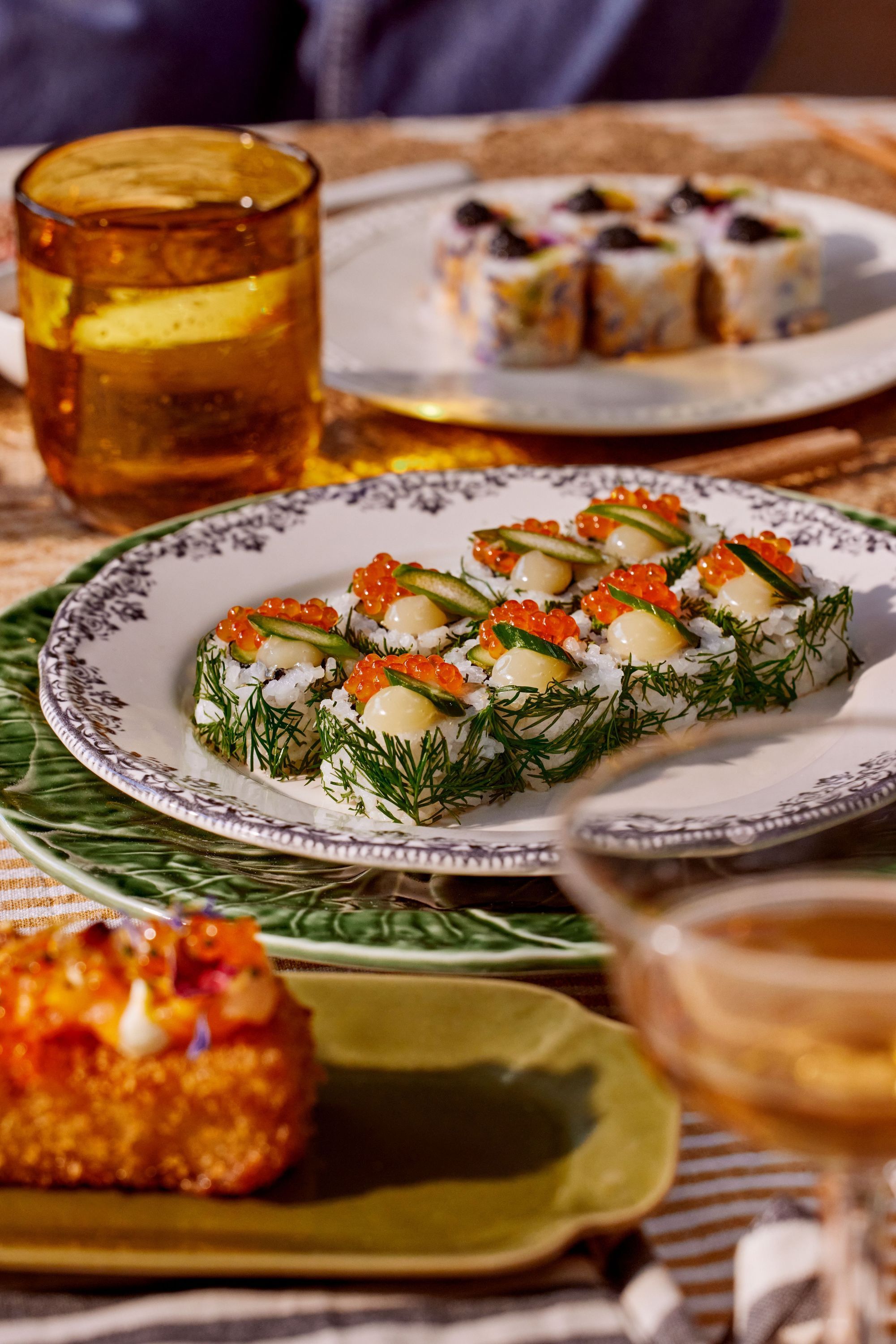 Sushis à emporter garnis d'œufs de poisson, crème et herbes sur assiette décorative avec verre ambre en arrière-plan.