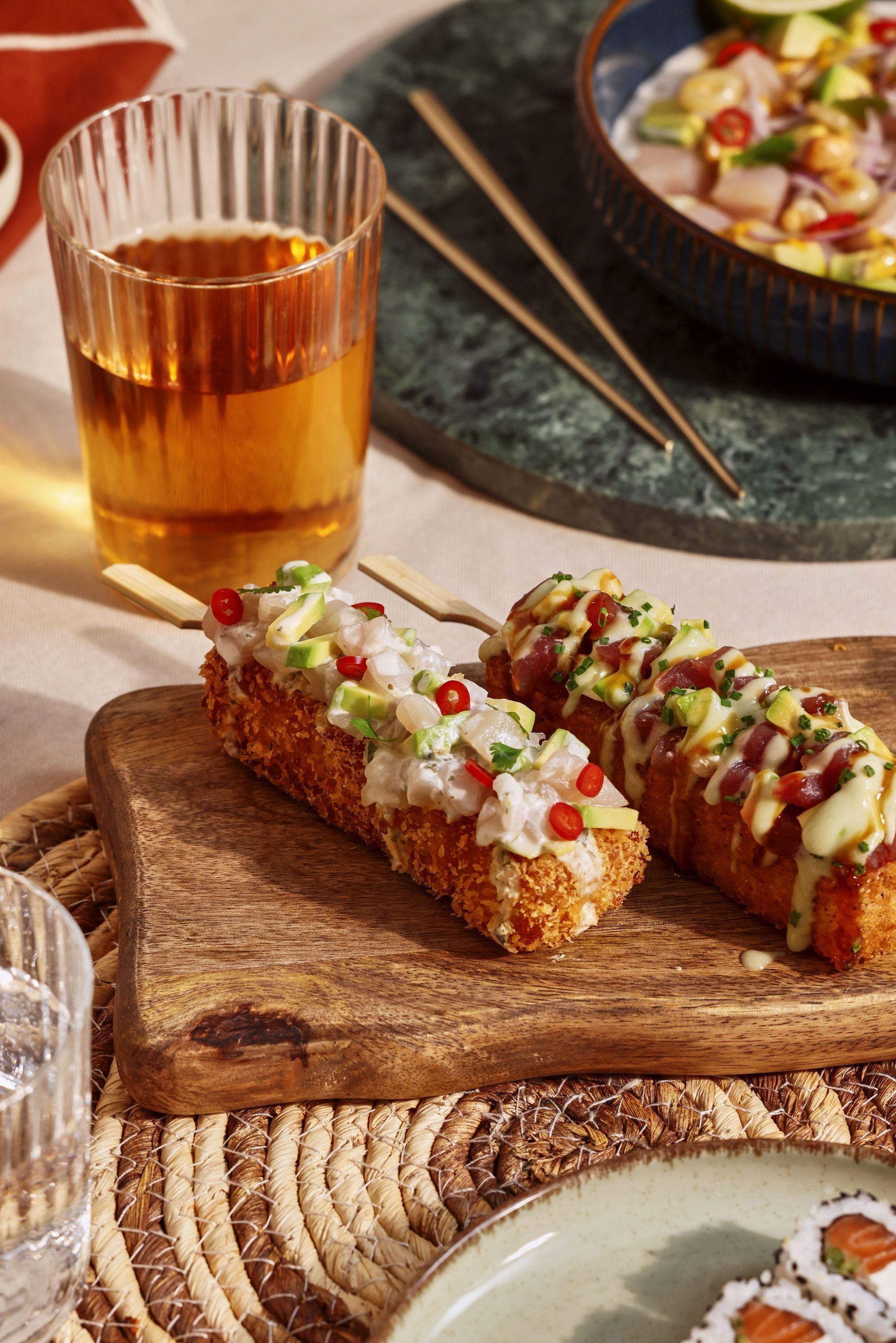 Sushis croustillants garnis de sauces et légumes, servis sur une planche en bois avec un verre de boisson à côté.
