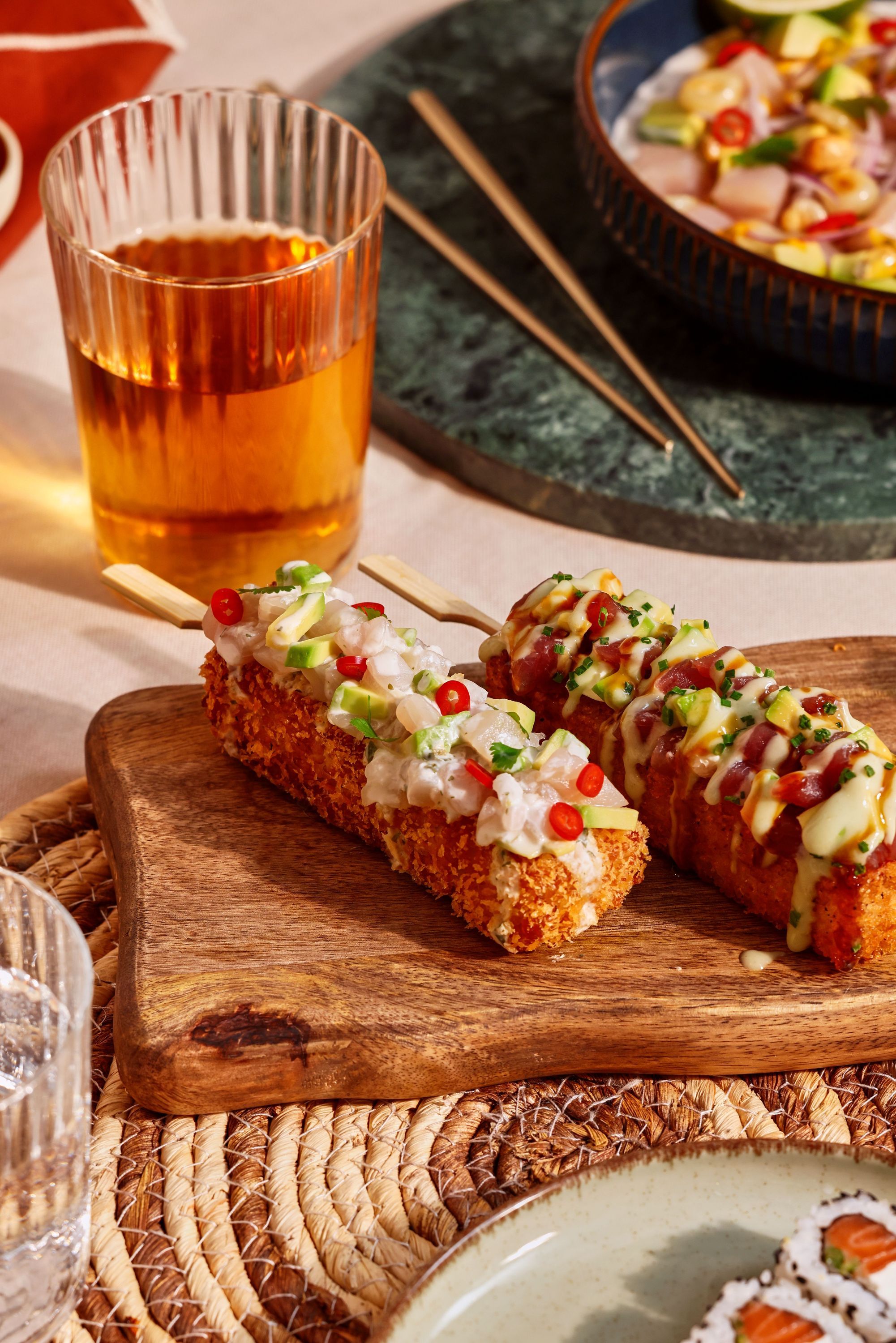 Sushis croustillants décorés d'oignons, piments et sauces, servis sur une planche en bois avec verre de boisson.