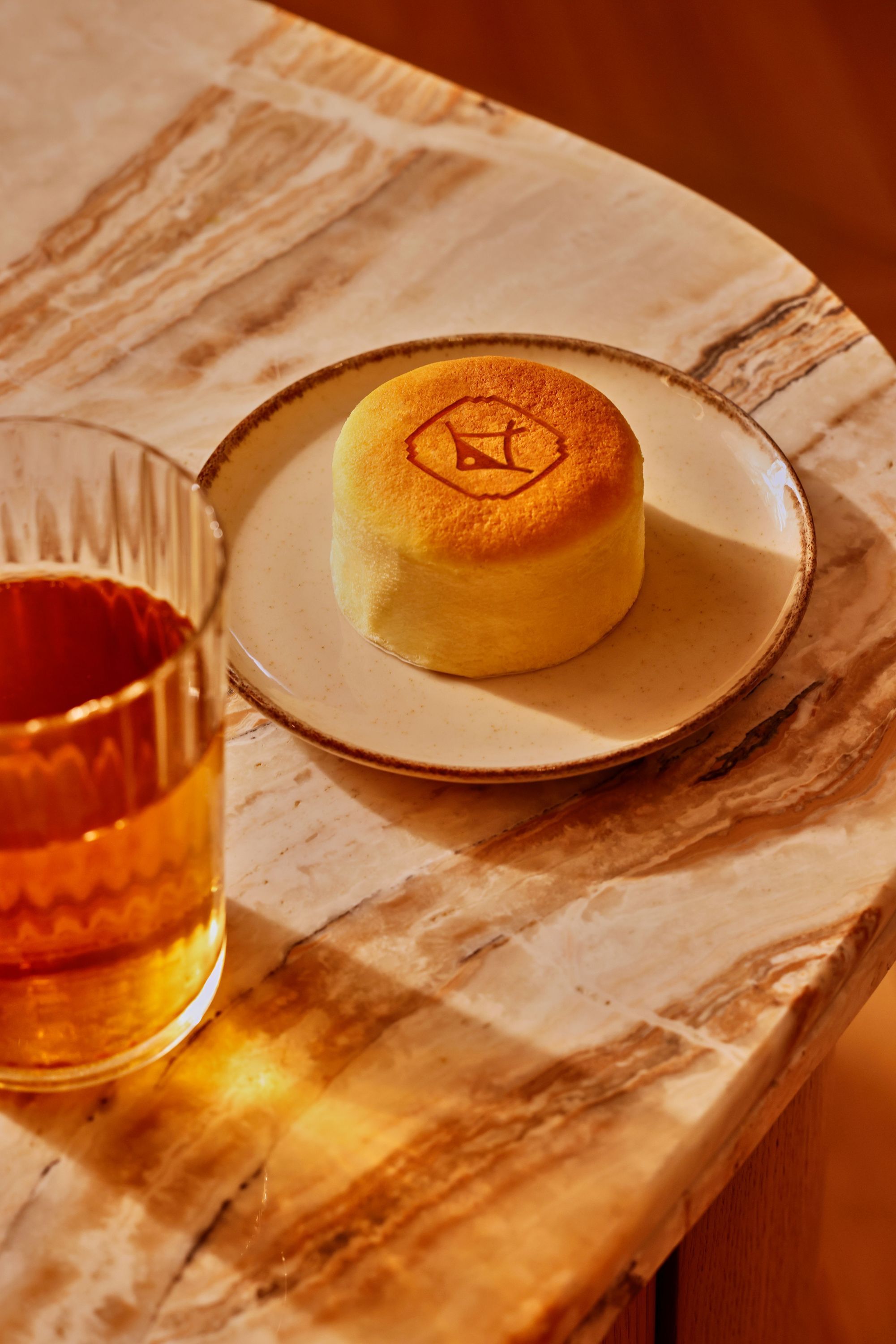 Petit gâteau japonais doré sur une assiette avec un symbole, posé sur une table en marbre clair à côté d'un verre de boisson.
